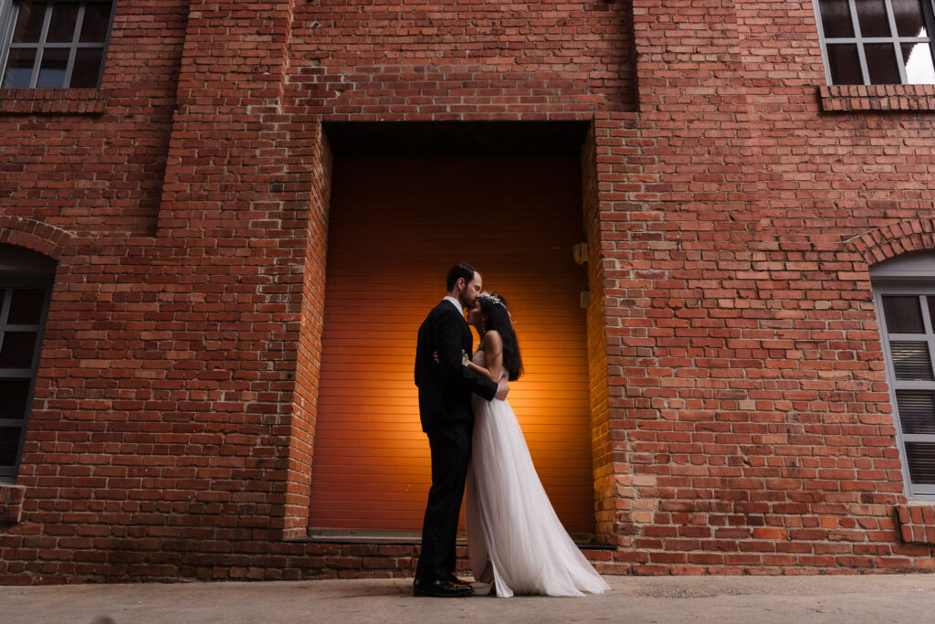 Bride and groom standing in front of a historic red brick wall at the American Tobacco Campus in Durham, NC, a popular spot for urban wedding photography near the Durham courthouse.